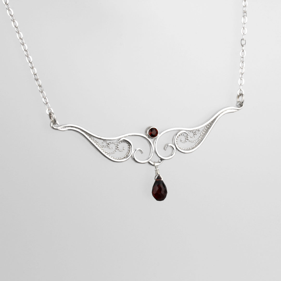 A graceful silver necklace with delicate filigree wirework, accented with deep red garnet, on a light background.