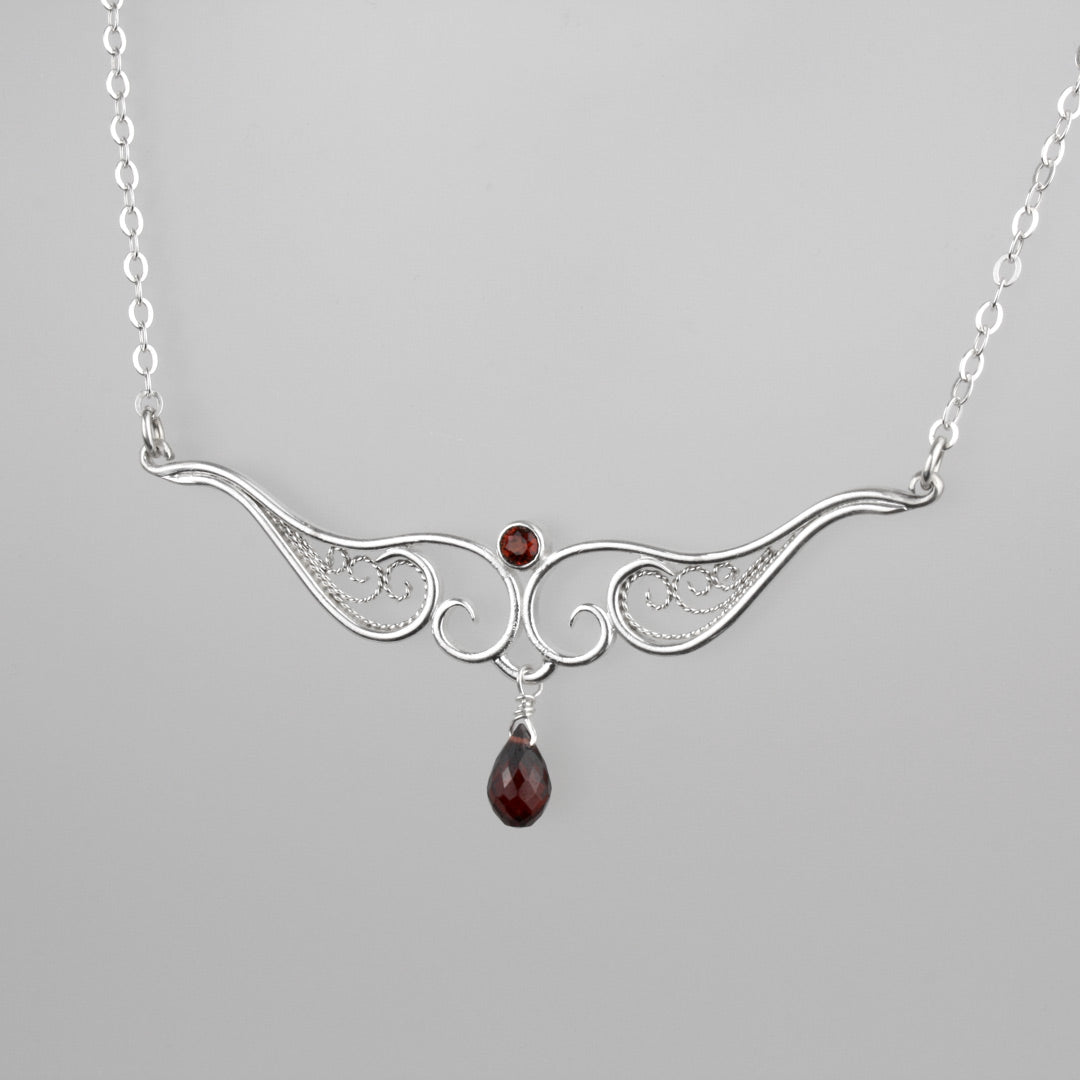 A graceful silver necklace with delicate filigree wirework, accented with deep red garnet, on a light background.