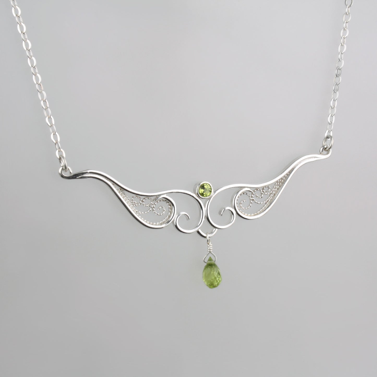 A graceful silver necklace with delicate filigree wirework, accented with green peridot, on a light background.