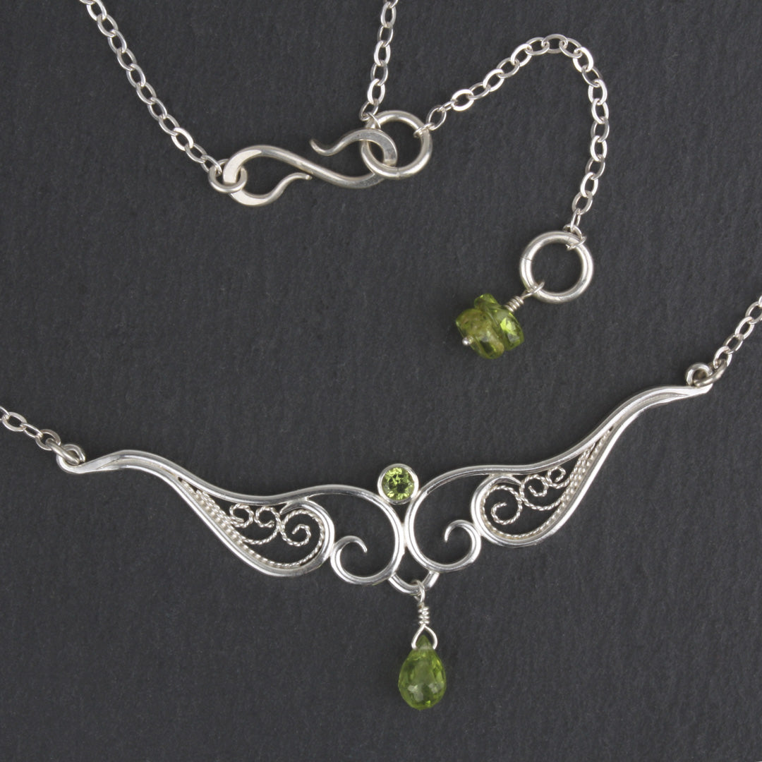 A graceful silver necklace with delicate filigree wirework, accented with green peridot, on a dark slate background.