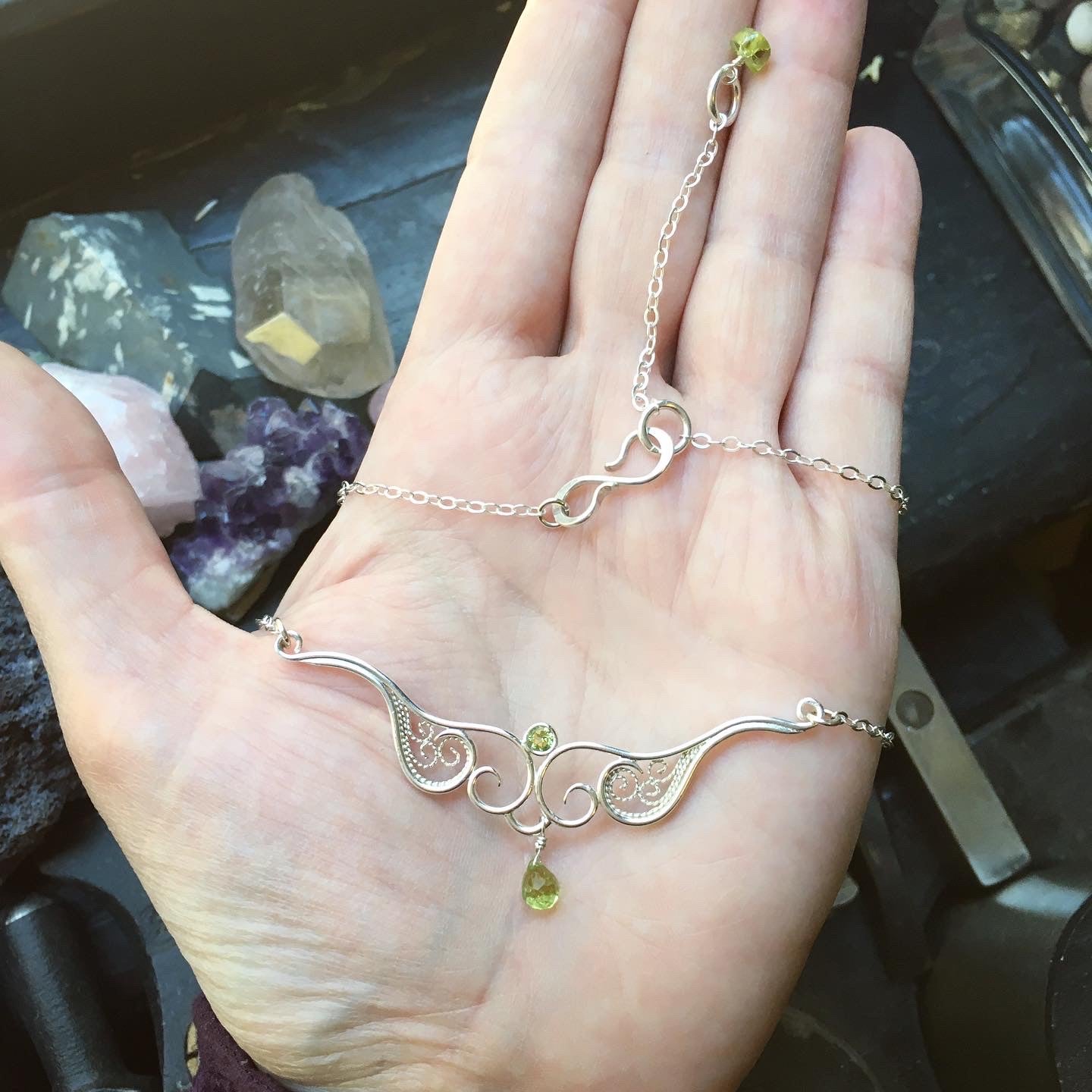 A graceful silver necklace with delicate filigree wirework, accented with green peridot, held in a hand.