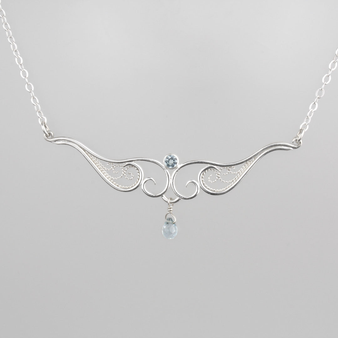 A graceful silver necklace with delicate filigree wirework, accented with sky blue topaz, on a light background.