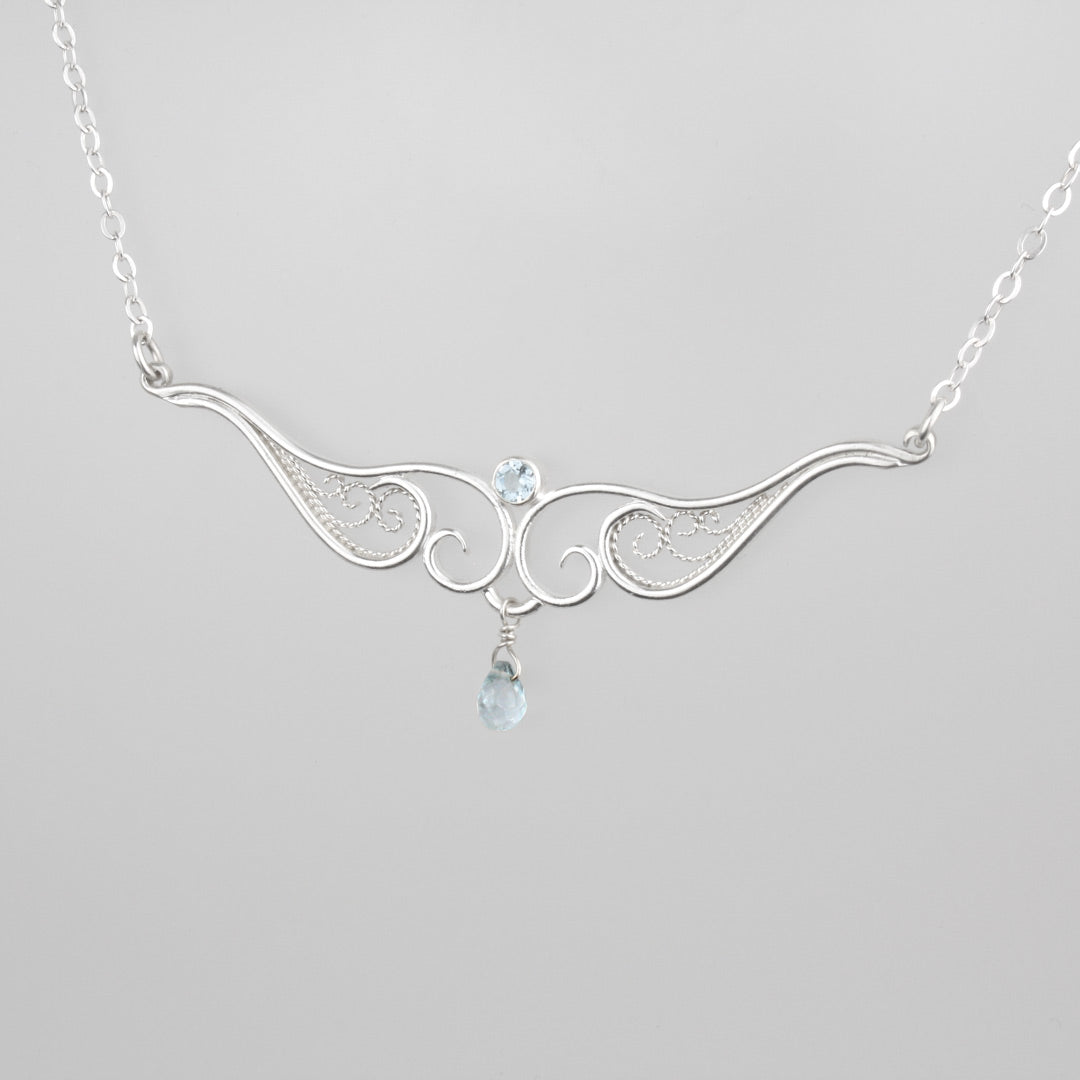 A graceful silver necklace with delicate filigree wirework, accented with sky blue topaz, on a light background.