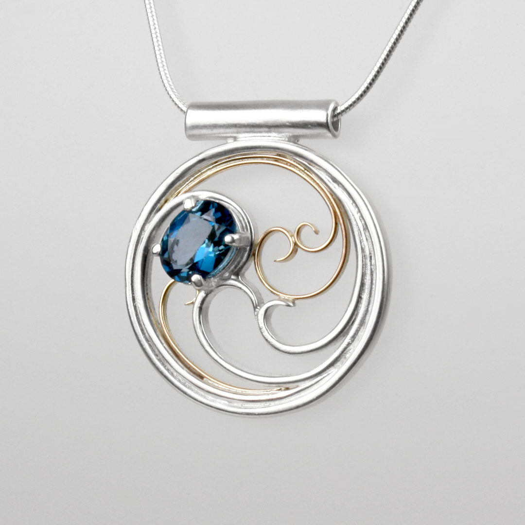 A circular pendant with cascading silver and gold waves encircling a deep blue oval topaz gem.