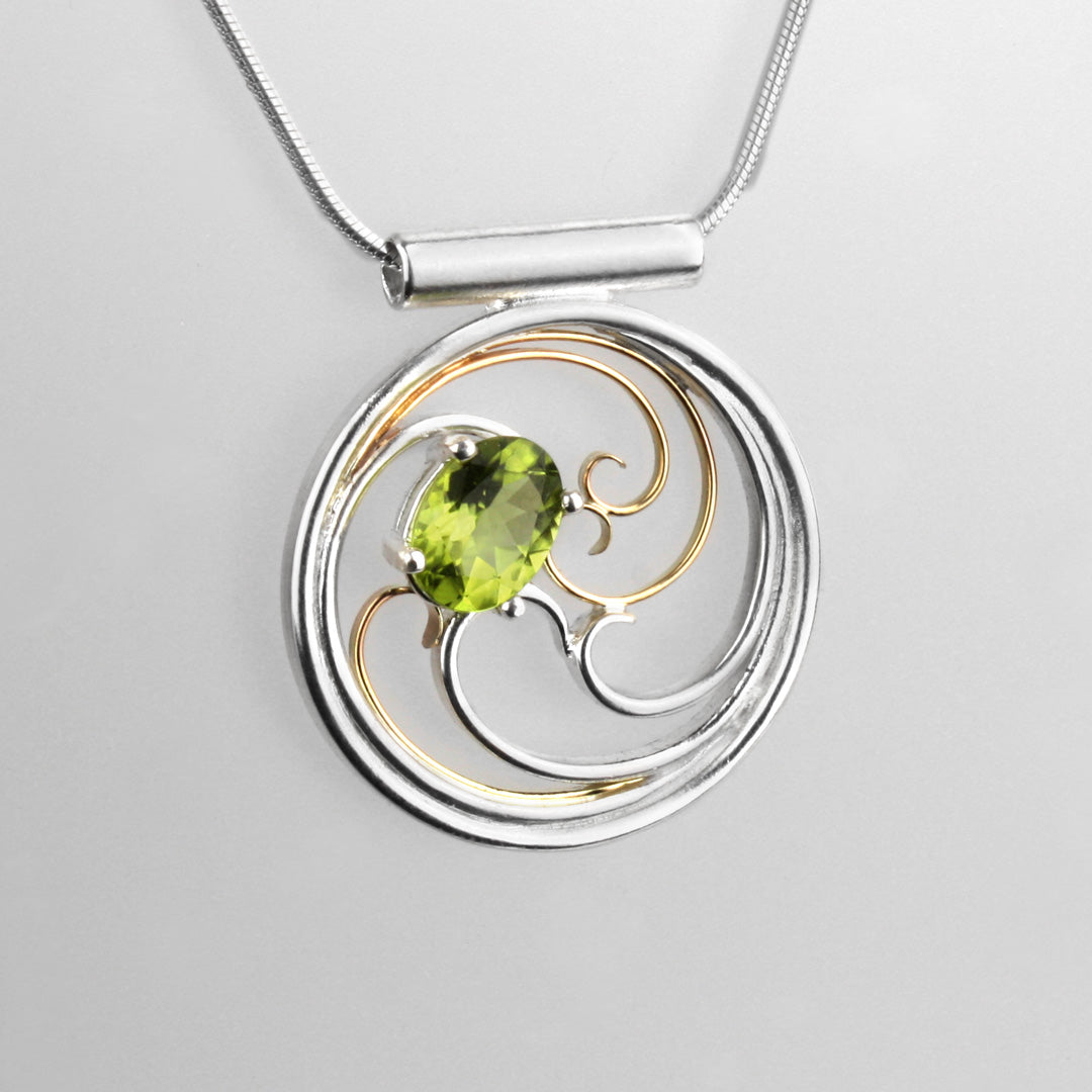 A circular pendant with cascading silver and gold waves encircling a brilliant green oval peridot gem.