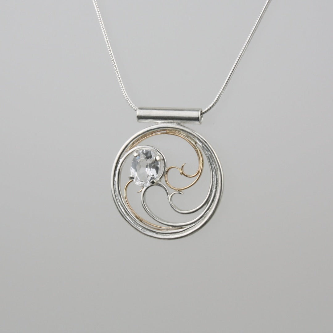 A circular pendant with cascading silver and gold waves encircling a sparkling white sapphire oval gem.