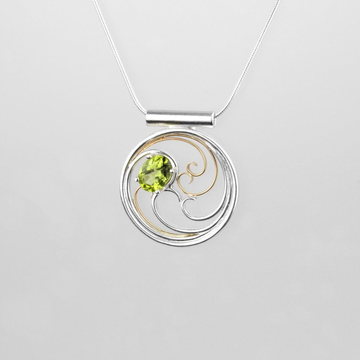 A circular pendant with cascading silver and gold waves encircling a brilliant green oval peridot gem.