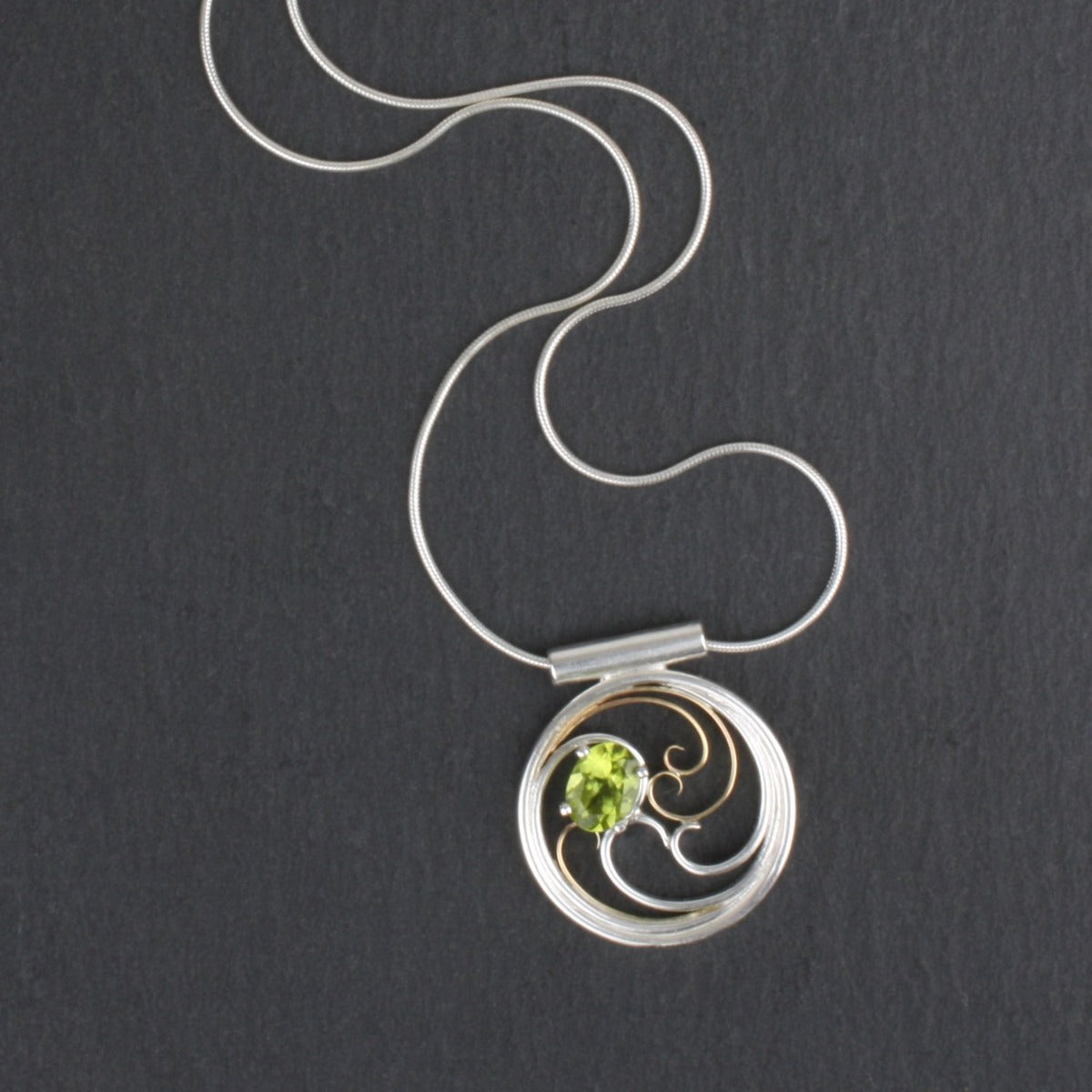 A circular pendant with cascading silver and gold waves encircling a brilliant green oval peridot gem.
