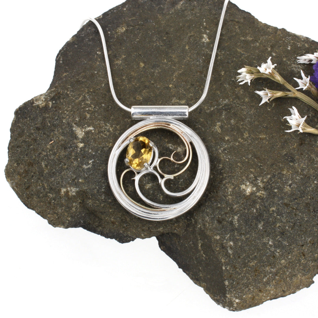 A circular pendant with cascading silver and gold waves encircling a golden yellow oval citrine gem.