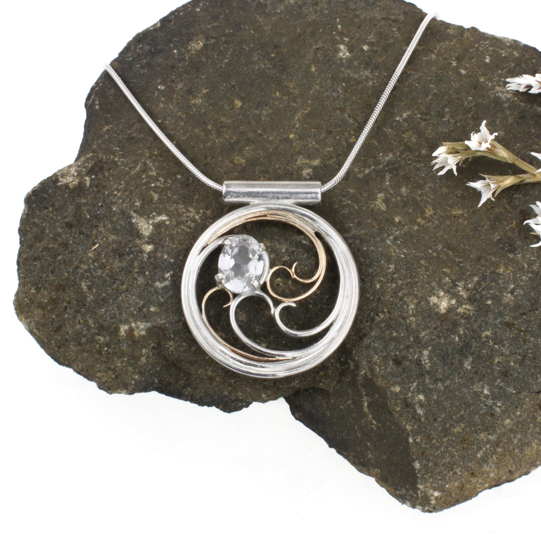 A circular pendant with cascading silver and gold waves encircling a sparkling white sapphire oval gem.