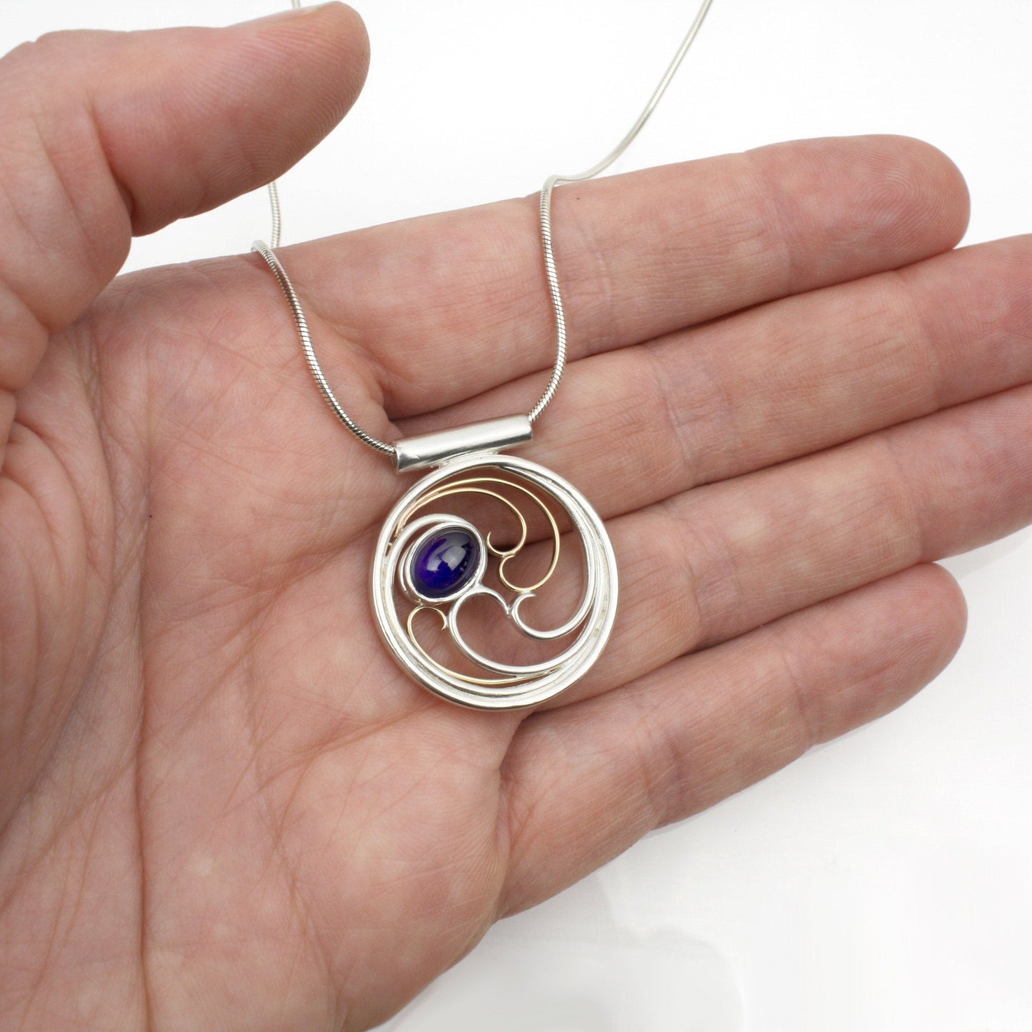 A circular pendant with cascading silver and gold waves encircling a deep indigo-violet oval cabochon iolite gem.