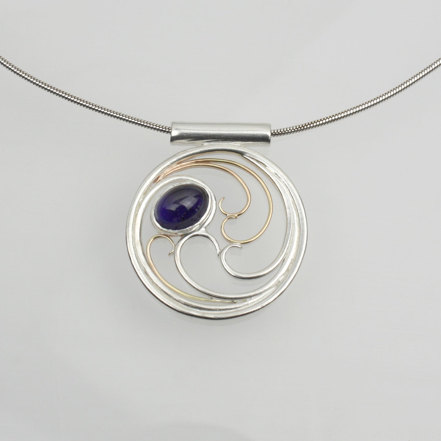A circular pendant with cascading silver and gold waves encircling a deep indigo-violet oval cabochon iolite gem.