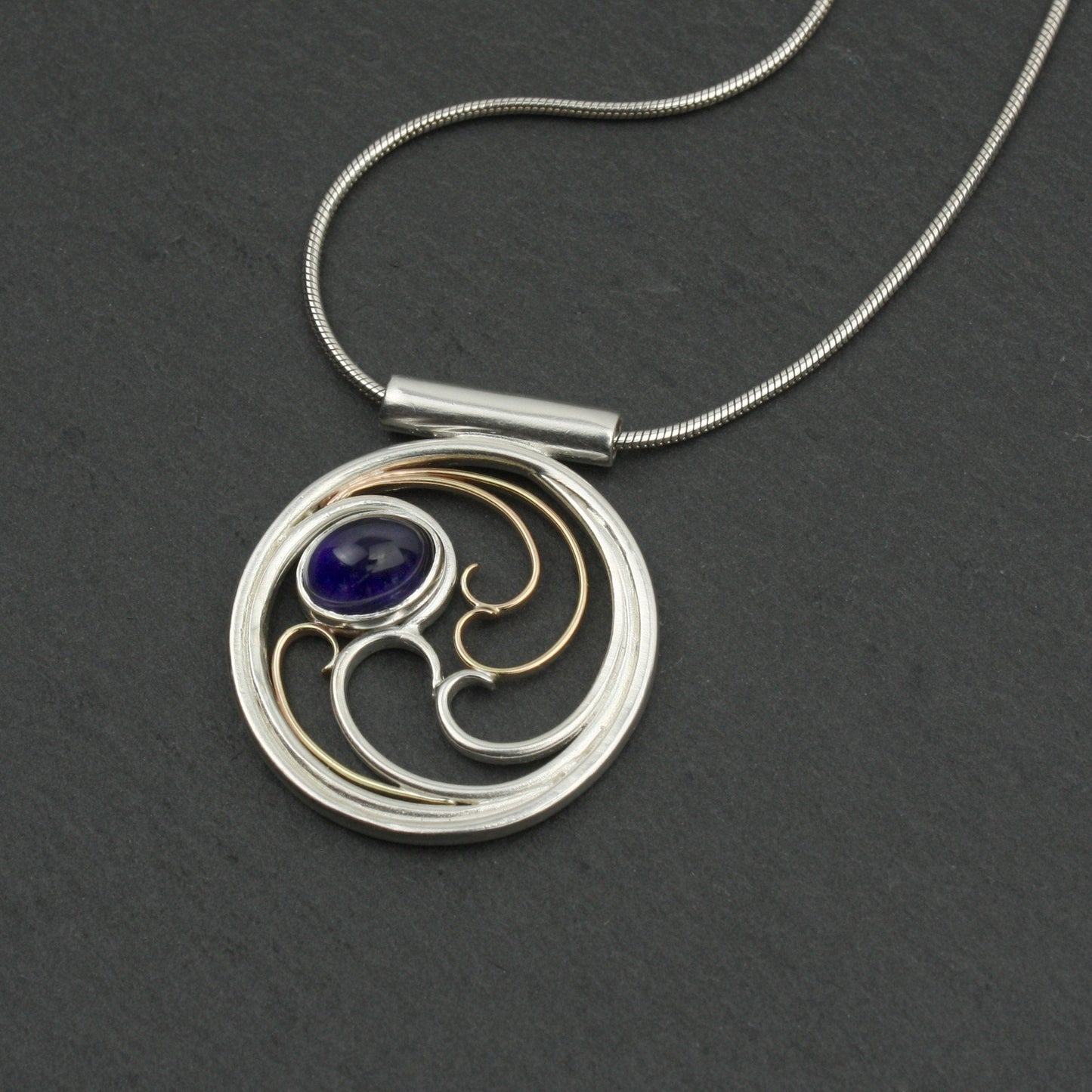 A circular pendant with cascading silver and gold waves encircling a deep indigo-violet oval cabochon iolite gem.