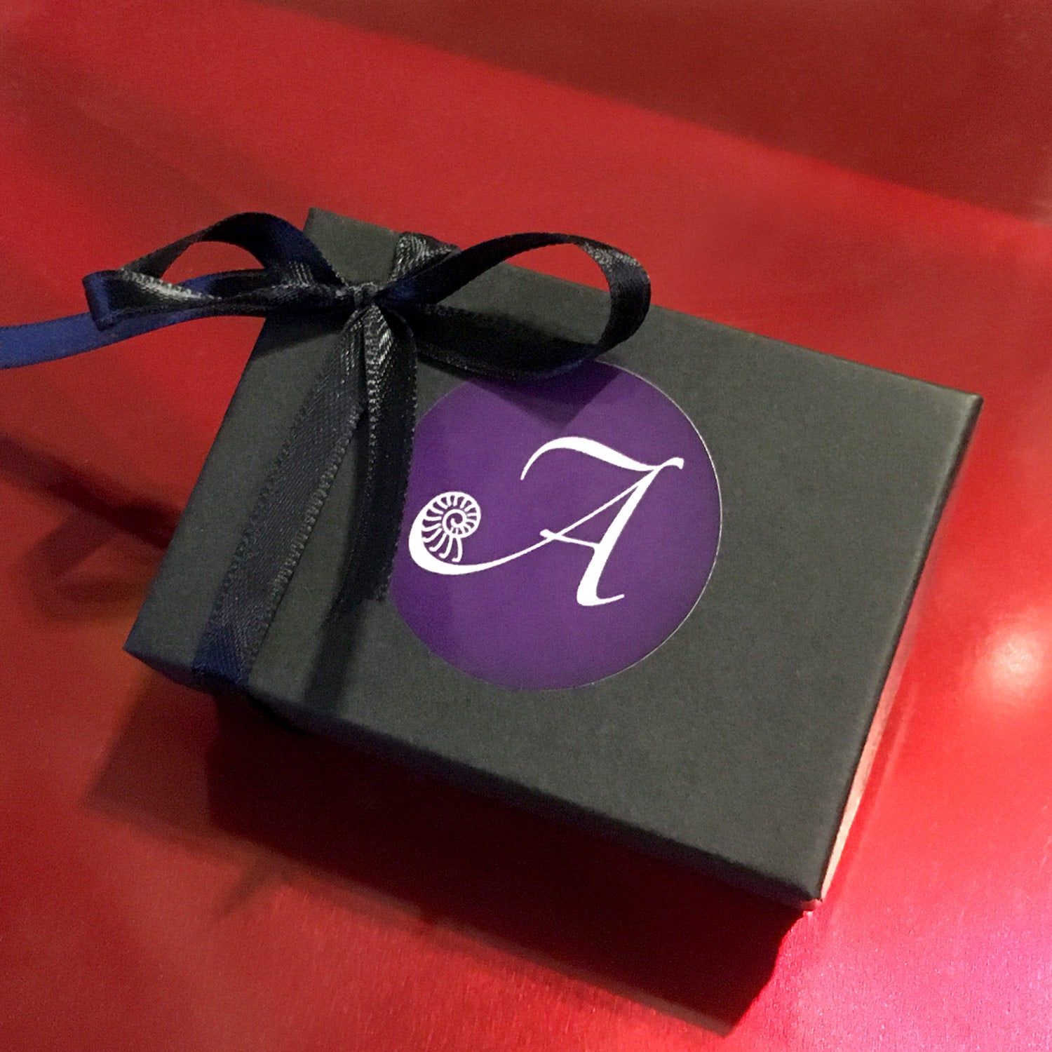 A black jewelry gift box with a purple label featuring the Arcana Metalwork logo, and a black satin bow.