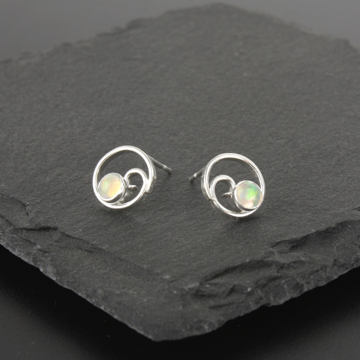 A photo of a pair of sterling silver post earrings with a spiral design, with green and orange iridescent opals, on a slate stone background.