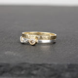 Two slender rings stacked on a slate stone surface. One ring is 14k yellow gold, with a tiny sparkling diamond (1mm size) embedded in the metal. The other ring has a larger, champagne-colored diamond on a sterling silver band, and the rings fit together. The gold ring has a hammered surface that catches the light.