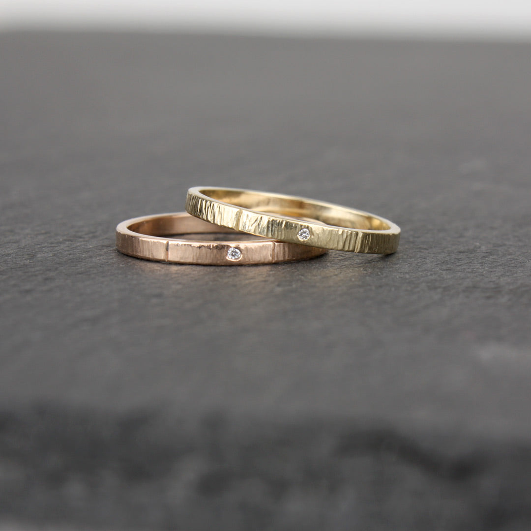 Two slender gold rings, one in 14k yellow gold, the other in 14k rose gold, on a slate stone. Each ring has a tiny sparkling diamond (1mm size) embedded in the metal. The rings have a hammered surface that catches the light.