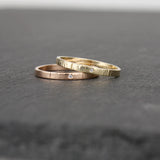 Two slender gold rings, one in 14k yellow gold, the other in 14k rose gold, on a slate stone. Each ring has a tiny sparkling diamond (1mm size) embedded in the metal. The rings have a hammered surface that catches the light.