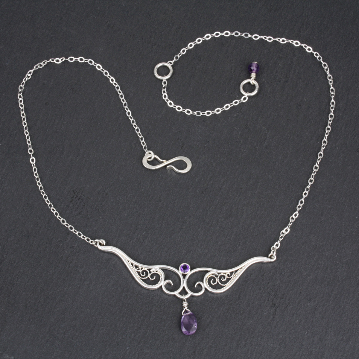 A graceful silver necklace with delicate filigree wirework, accented with purple amethyst, on a slate stone background.