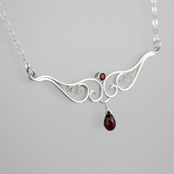 A graceful silver necklace with delicate filigree wirework, accented with deep red garnet, on a light background.