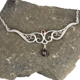 A graceful silver necklace with delicate filigree wirework, accented with deep red garnet, on a stone background.