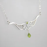 A graceful silver necklace with delicate filigree wirework, accented with green peridot, on a light background.