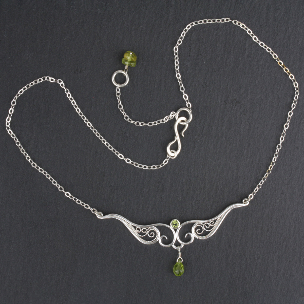 A graceful silver necklace with delicate filigree wirework, accented with green peridot, on a dark slate background.