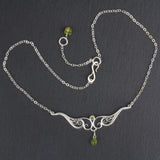 A graceful silver necklace with delicate filigree wirework, accented with green peridot, on a dark slate background.