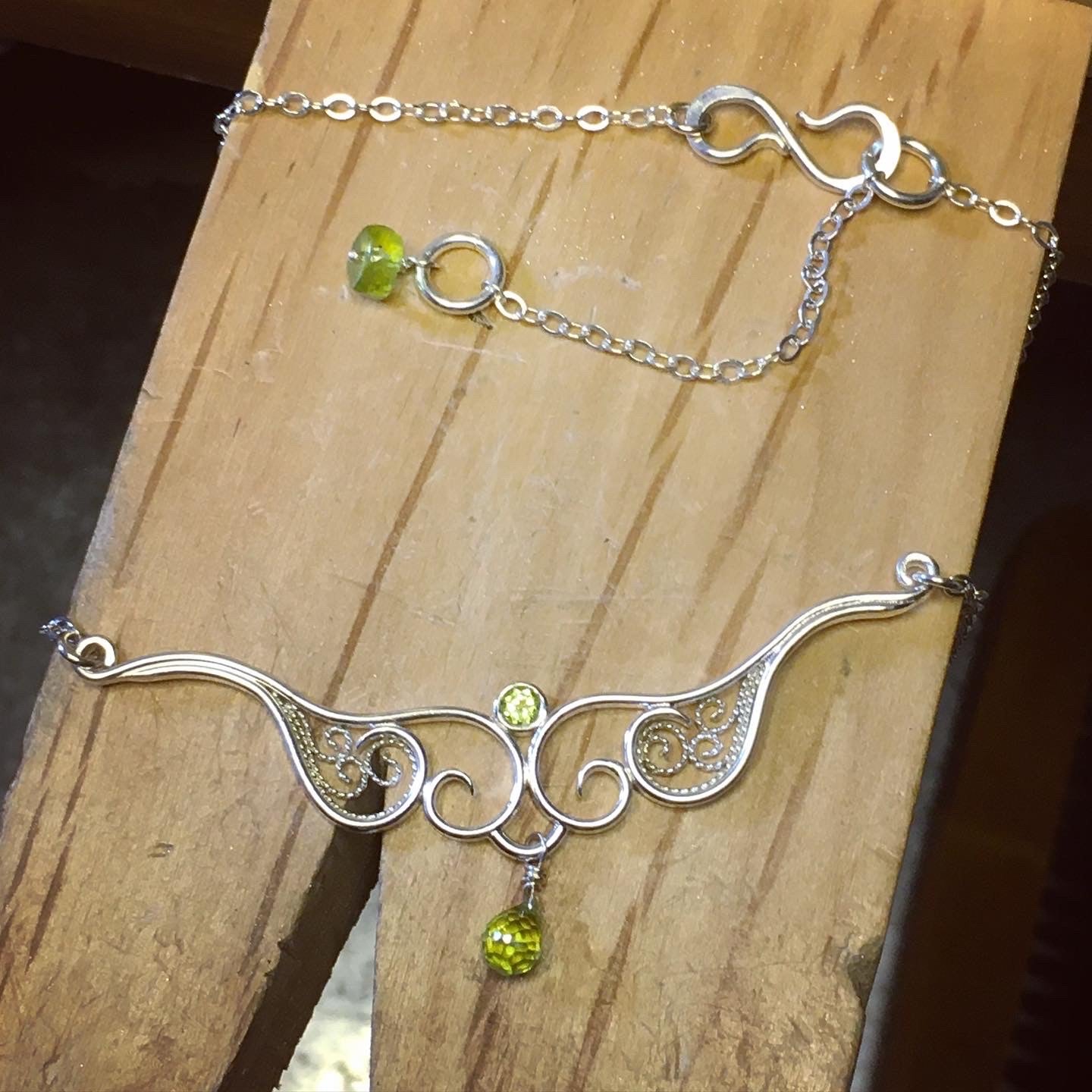 A graceful silver necklace with delicate filigree wirework, accented with green peridot, on a wooden jeweler's bench pin.