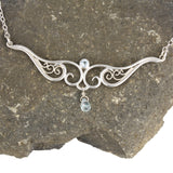 A graceful silver necklace with delicate filigree wirework, accented with sky blue topaz, on a stone background.