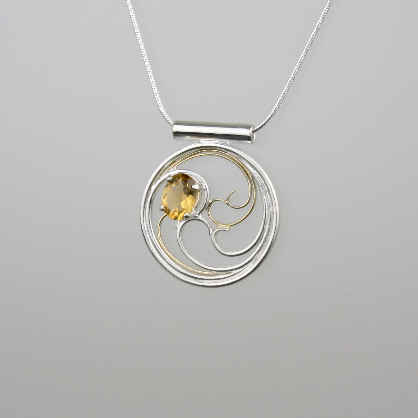A circular pendant with cascading silver and gold waves encircling a golden yellow oval citrine gem.