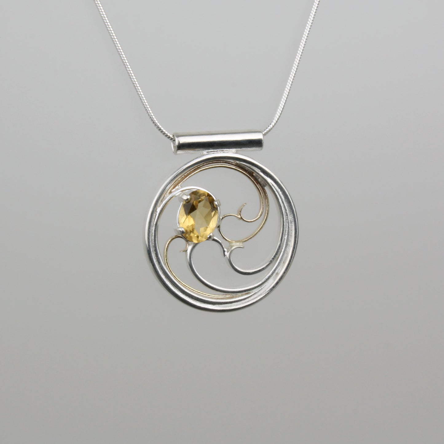 A circular pendant with cascading silver and gold waves encircling a golden yellow oval citrine gem.