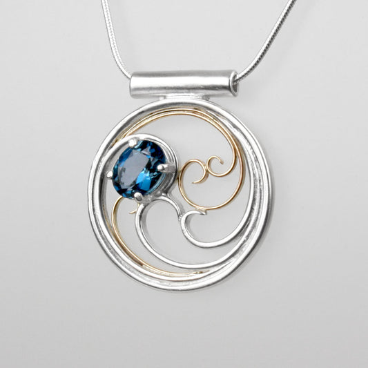 A circular pendant with cascading silver and gold waves encircling a deep blue oval topaz gem.