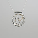 A circular pendant with cascading silver and gold waves encircling a sparkling white sapphire oval gem.