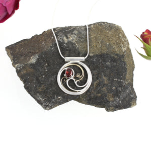 A circular pendant with cascading silver and gold waves encircling a deep red oval garnet gem.