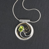 A circular pendant with cascading silver and gold waves encircling a brilliant green oval peridot gem.