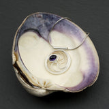 A circular pendant with cascading silver and gold waves encircling a deep indigo-violet oval cabochon iolite gem.